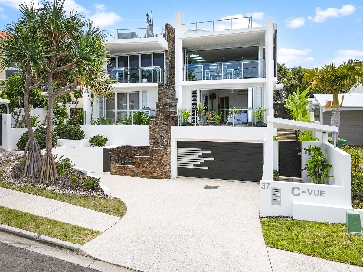 The exterior of a modern home is displayed, featuring clean lines and a combination of stone and white walls. Lush greenery surrounds the property, enhancing the contemporary design. A driveway leads to a garage with a sleek door, inviting guests to explore further.