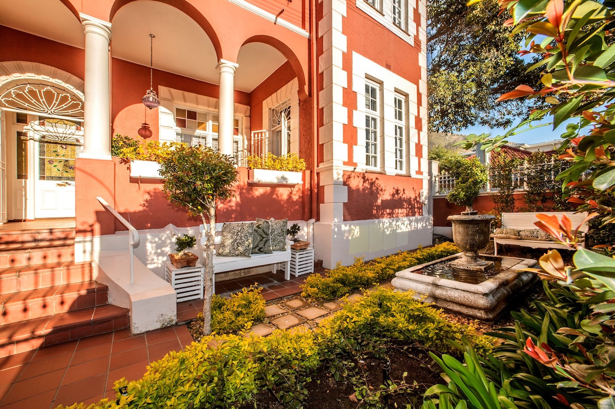 The exterior features a charming red and white building with arched columns and large windows. A well-maintained garden surrounds a small fountain, while a bench sits on the porch, inviting relaxation amidst vibrant greenery.