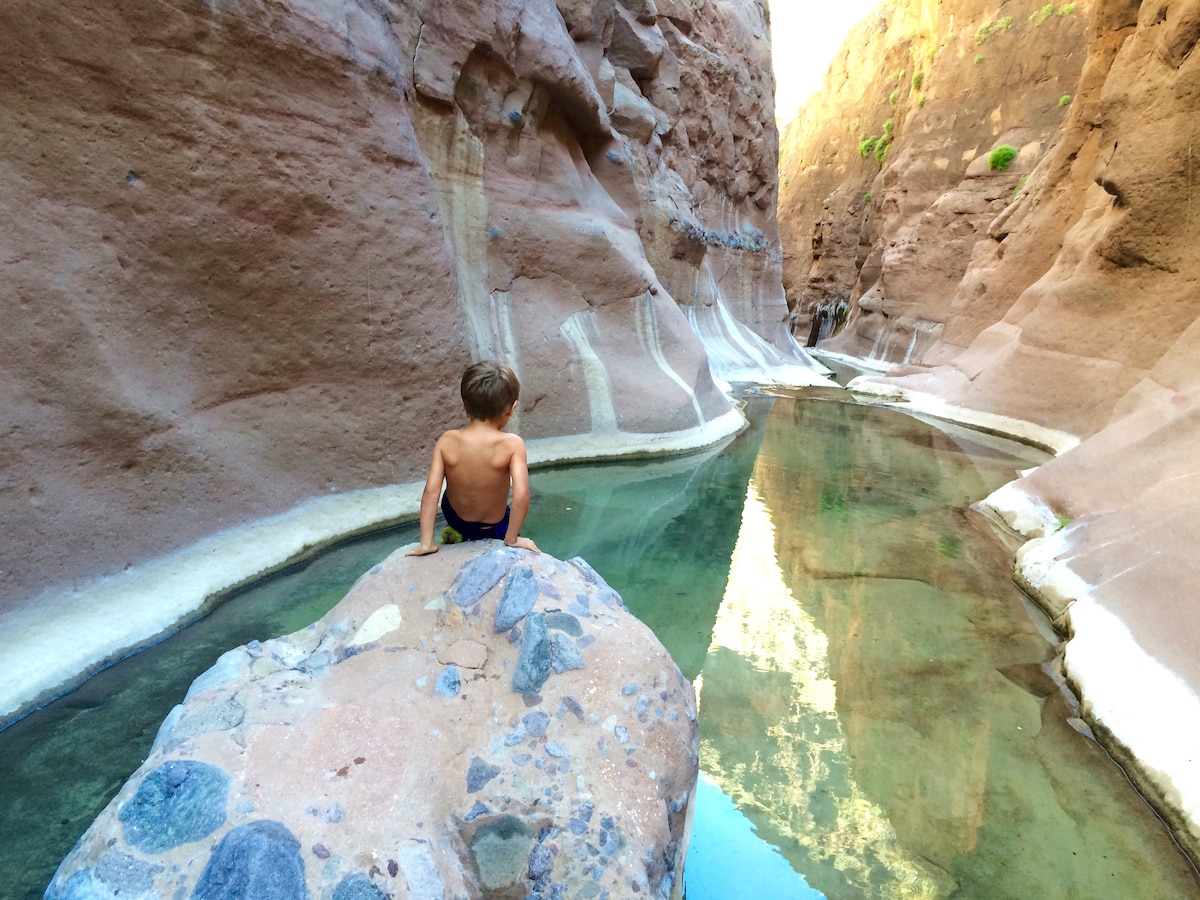 A child sits on a smooth rock by a calm waterway, flanked by towering, reddish-brown canyon walls. The still water reflects the rugged landscape and the warm light filtering through the canyon. Vegetation is visible atop the cliffs in the background.
