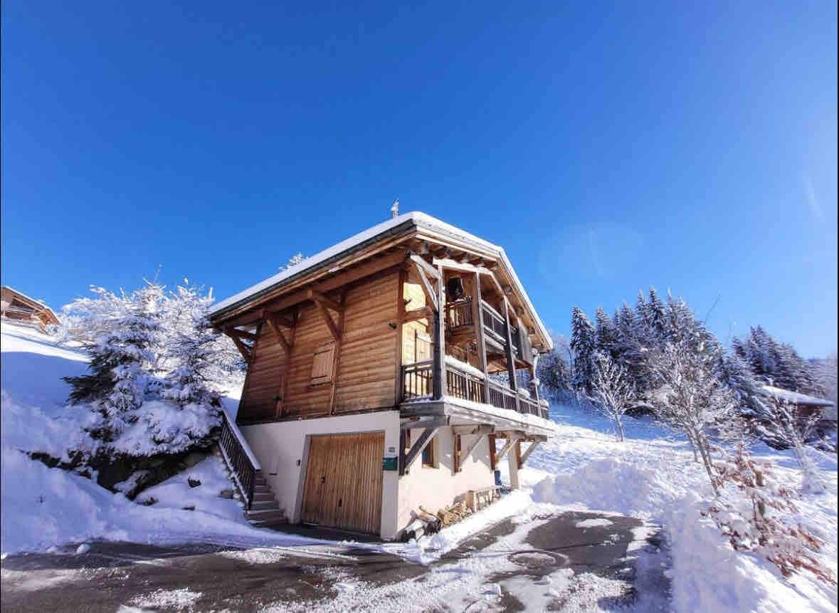 A chalet with a wooden exterior is positioned against a backdrop of snow-covered ground and evergreen trees. The structure features a balcony with railings, and a set of stairs leads to the entrance. Bright blue sky enhances the serene winter landscape.