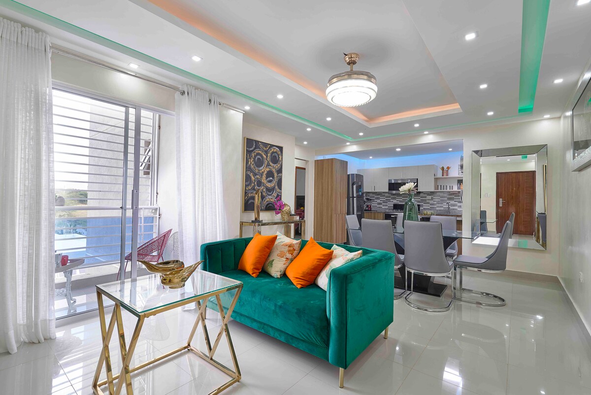 A modern living space features a teal sofa adorned with vibrant orange cushions. A glass coffee table sits in front of the sofa, while a dining area with grey chairs and a sleek kitchen is visible in the background. Natural light filters through sheer curtains.