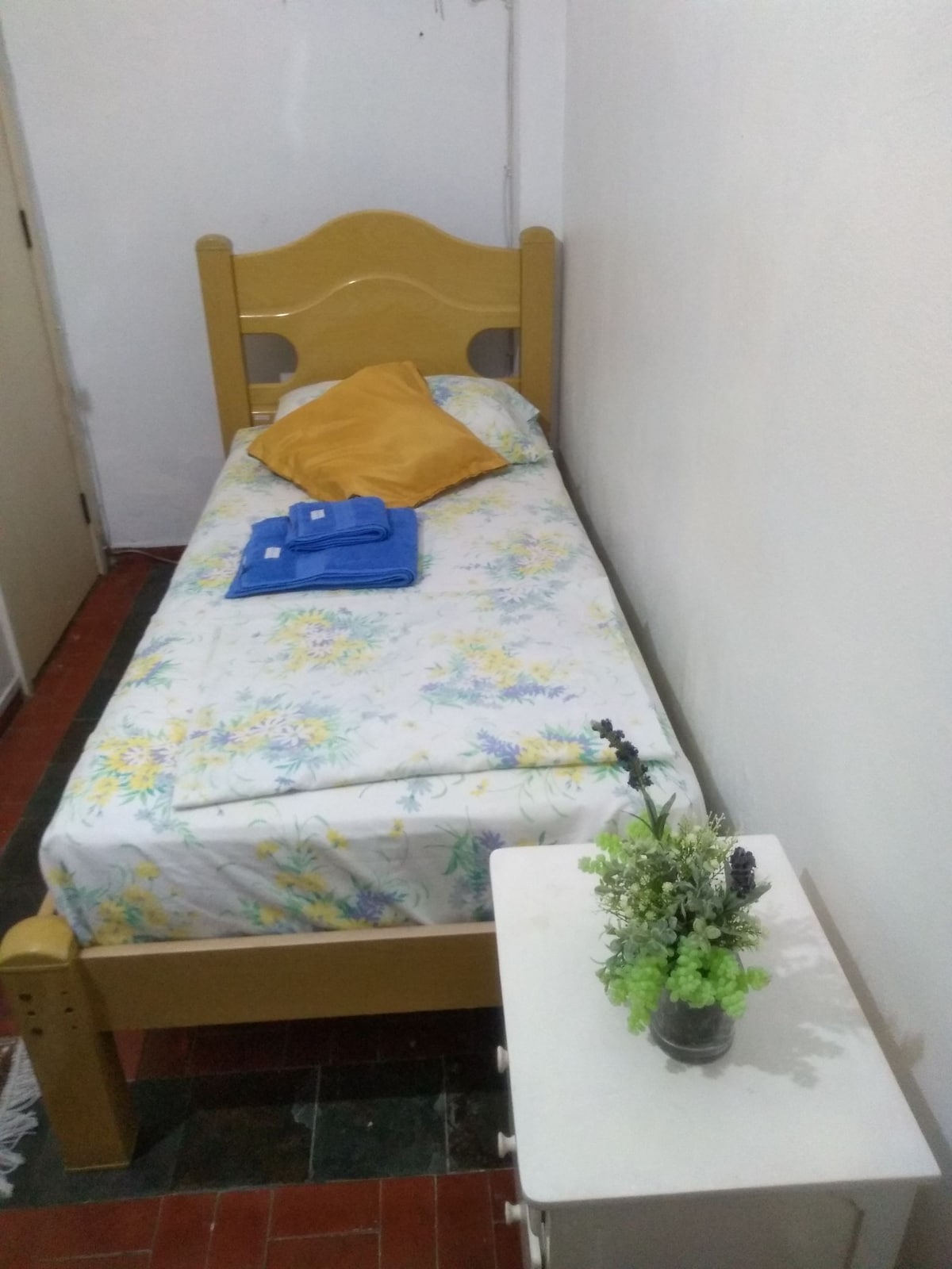 A single bed is dressed with a floral-patterned sheet, complemented by a yellow pillow and folded towels in blue. Nearby, a small nightstand holds a simple vase with a selection of artificial greenery, enhancing the practical layout of the space.