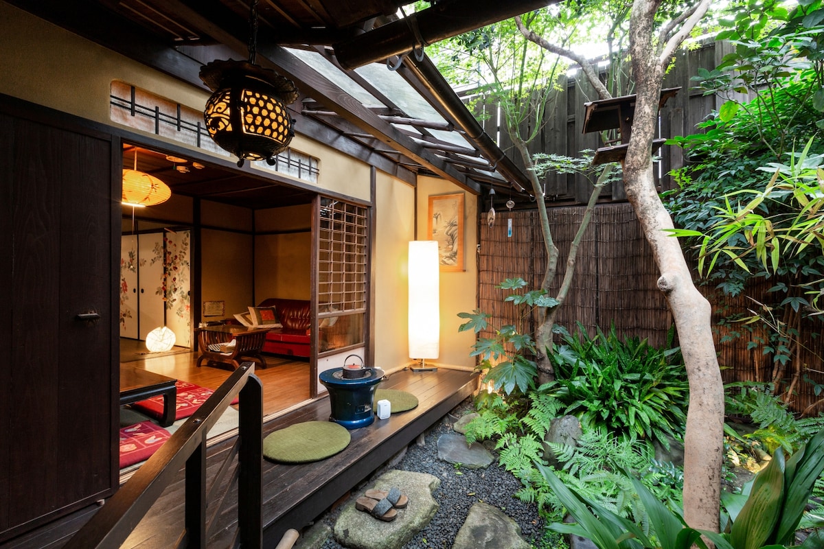 A charming entrance leads to a serene garden, framed by lush greenery and smooth stones. Soft lighting illuminates the space, highlighting traditional architectural details. The wooden pathway connects the indoor area to the tranquil outdoors, inviting a peaceful atmosphere.