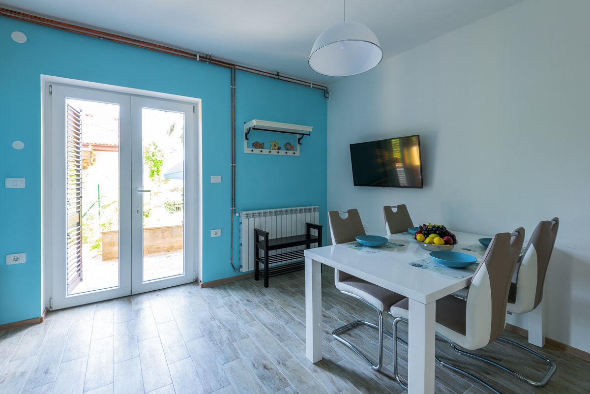 A dining area is presented with a white table and four beige chairs. A flat-screen TV is mounted on the wall, and a bowl of fruit is placed at the center of the table. Light enters through double glass doors, which lead to the outdoor space.