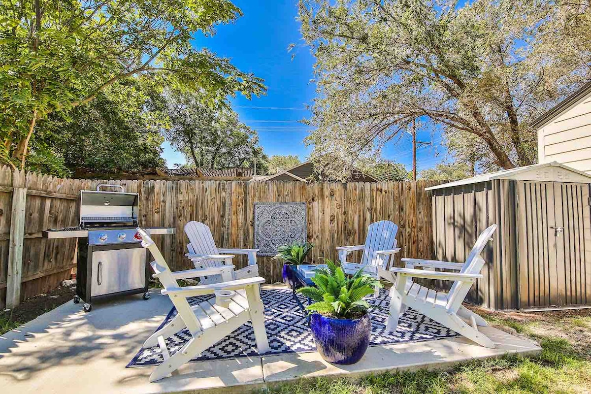 A fenced backyard area features a grilling station, surrounded by four adirondack chairs positioned on a patterned outdoor rug. Lush green plants are placed in an oversized pot, enhancing the serene outdoor setting with a wooden storage shed visible in the background.