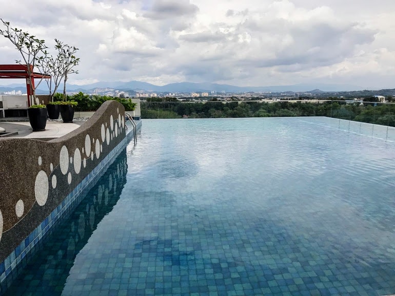 An infinity pool is visible, surrounded by a decorative edge that features circular motifs. The calm water reflects the sky and nearby greenery, with a distant view of rolling hills and cityscapes under a partly cloudy sky.