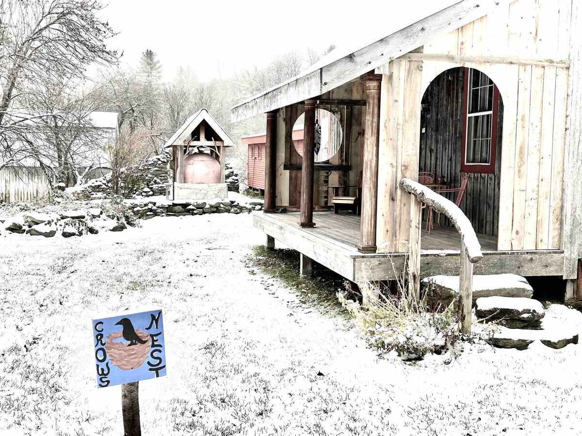 The exterior of Crow's Nest is presented in a snowy landscape, featuring a wooden deck with arches and a welcoming porch. A whimsical sign indicates the property name, with texture and detail visible in the surrounding winter scenery.