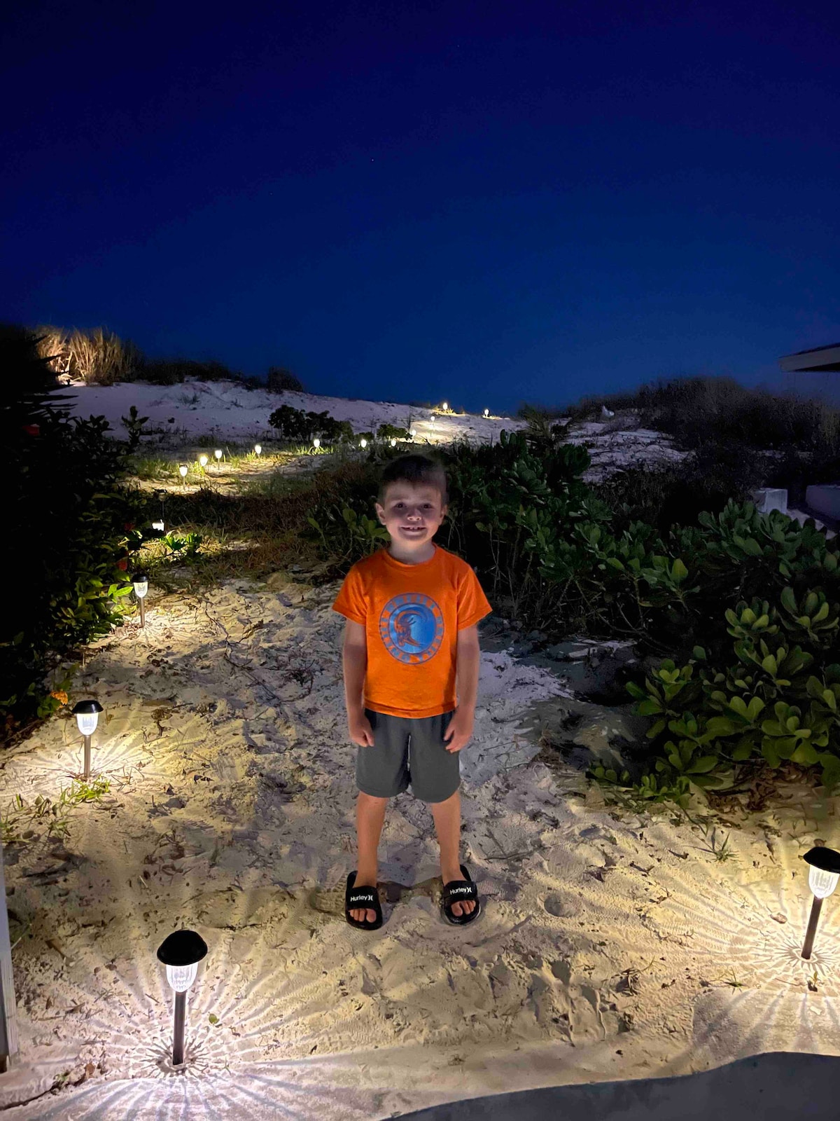 A softly illuminated path meanders through a sandy area, lined with small lights that create a warm ambiance. Low vegetation flanks the walkway, with a backdrop of gentle sand dunes under a night sky.