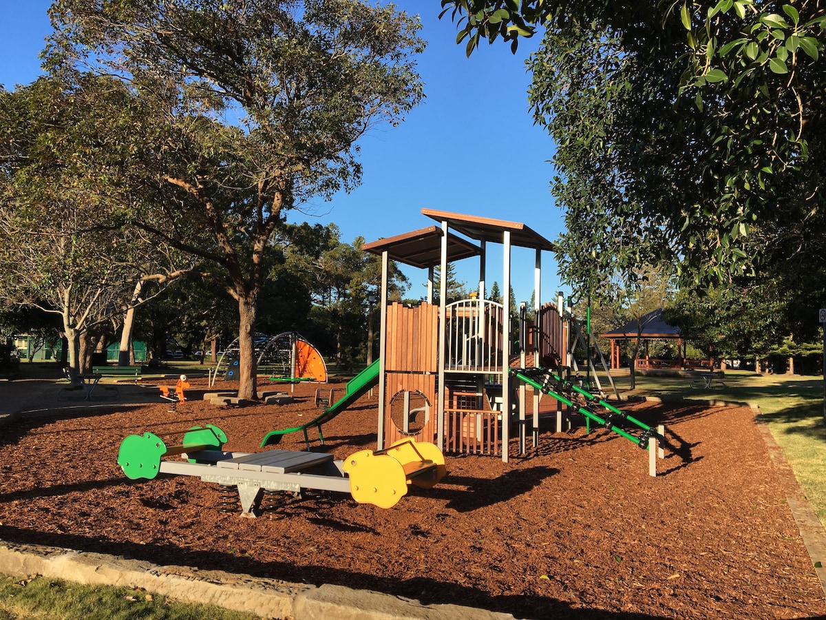 A well-maintained playground is set within a spacious area, featuring a wooden climbing structure with slides and ramps. Surrounding the play equipment, soft mulch provides a safe landing, while shaded trees contribute to a pleasant outdoor environment.