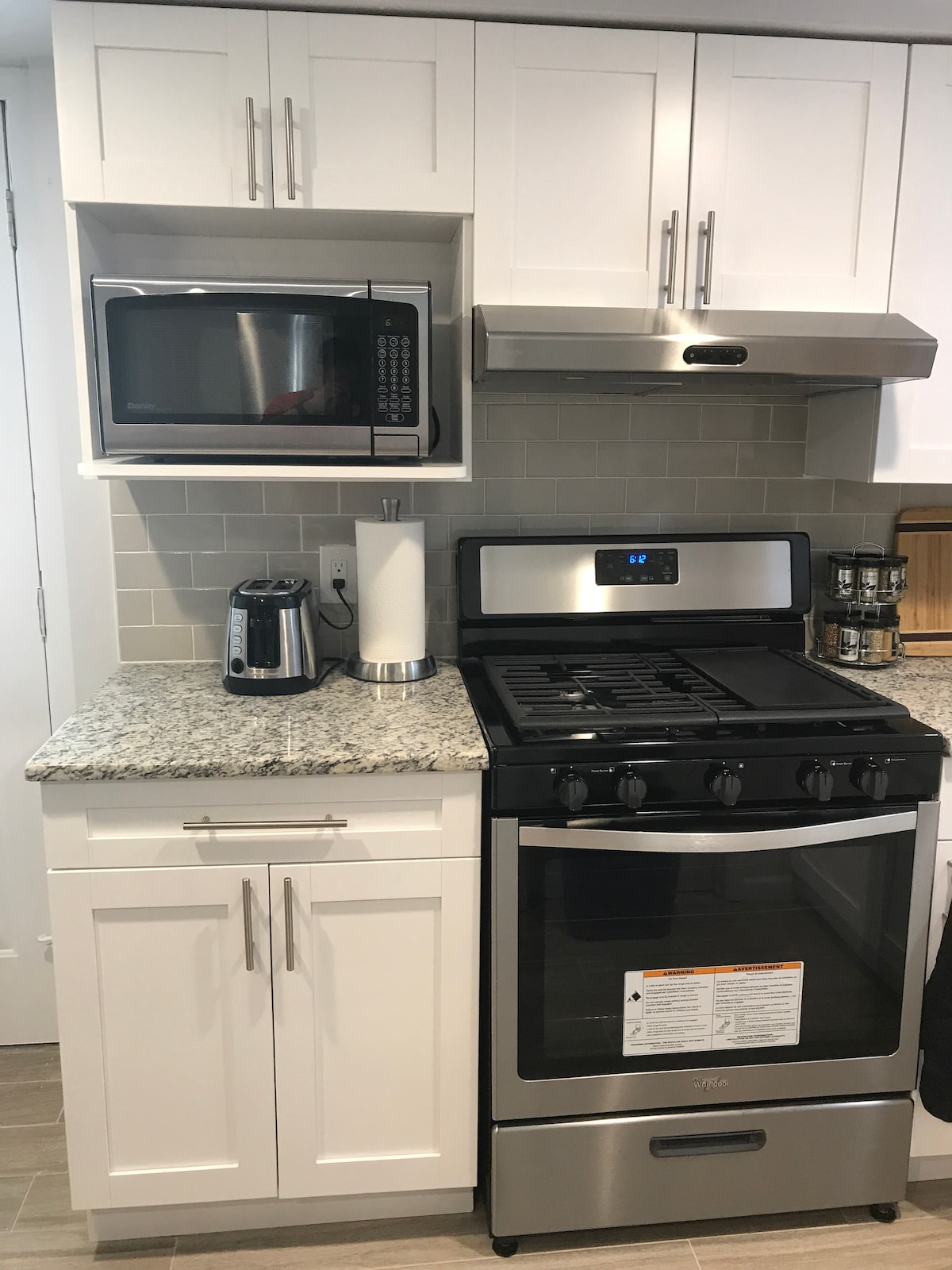 A modern kitchen is displayed, featuring stainless steel appliances, including a gas stove, microwave, and coffee maker. Light-colored cabinetry complements the granite countertop. A paper towel holder and a spice rack add functional elements to the well-organized space.