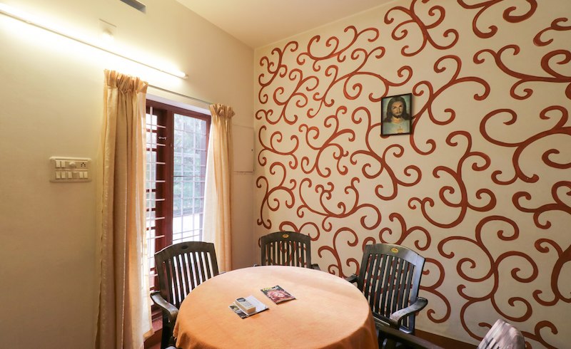 A cozy dining area is featured with a round table covered by an orange tablecloth. Four dark chairs surround the table, and a decorative wall adorned with swirls adds character. Natural light enters through a window with sheer curtains, enhancing the inviting space.