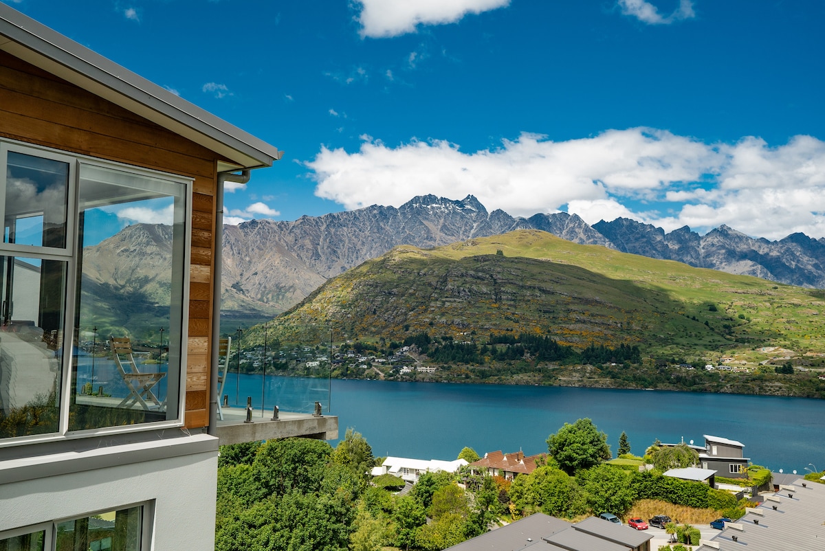 The image depicts a modern house positioned on a hill, featuring large windows that provide expansive views of the mountains and Lake Wakatipu. Clear blue skies complement the lush greenery below, creating a serene backdrop to the tranquil setting.