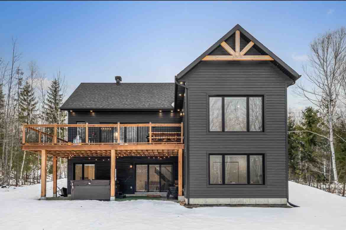 A modern chalet with a dark exterior is surrounded by snow-covered terrain. A spacious wooden deck extends from the upper level, featuring outdoor seating. Large windows provide ample natural light to the structure, which is set against a backdrop of tall trees.