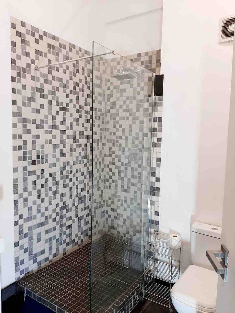 A modern bathroom features a glass shower enclosure with a mosaic tile accent wall. A sleek toilet is positioned nearby, and a small metal cart holds essentials. Clean lines and minimalistic design contribute to a tidy atmosphere.