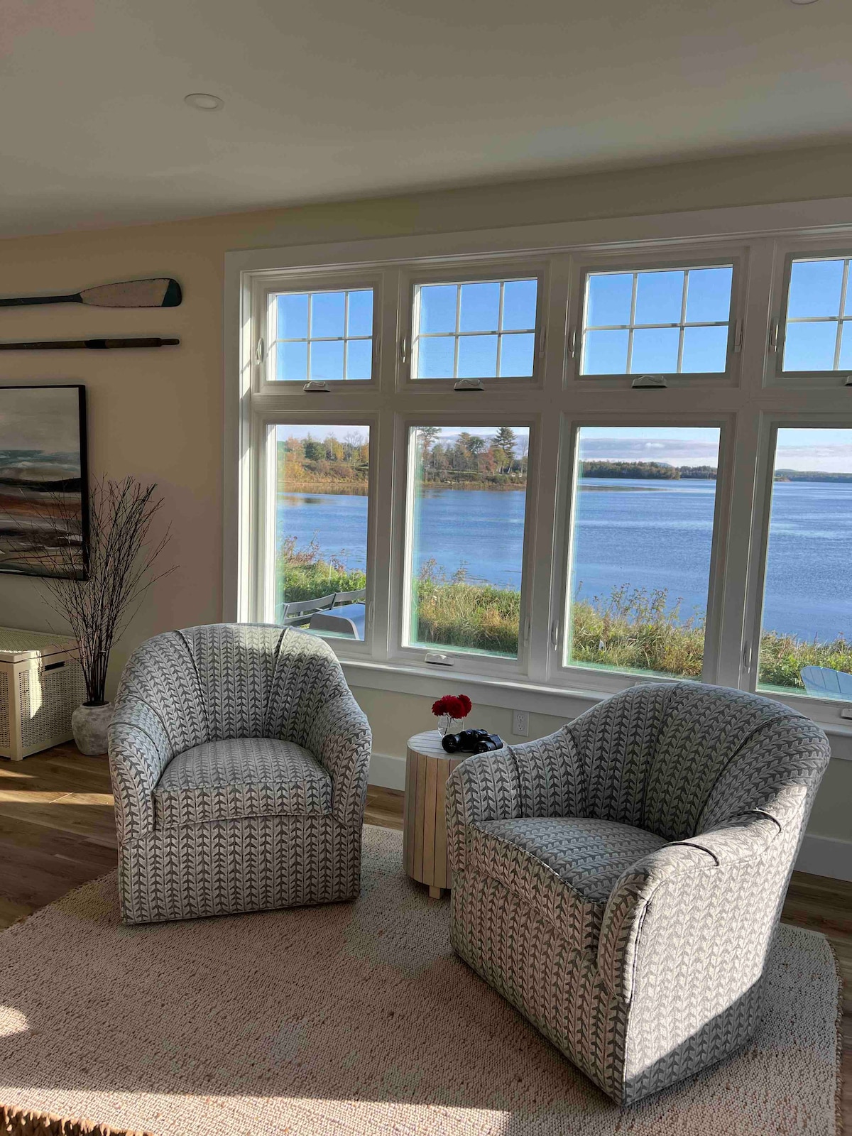 Two comfortable chairs are positioned near a large window, offering unobstructed views of the water and surrounding landscape. The space is illuminated by natural light, and a small table between the chairs holds a decorative item, enhancing the serene atmosphere.