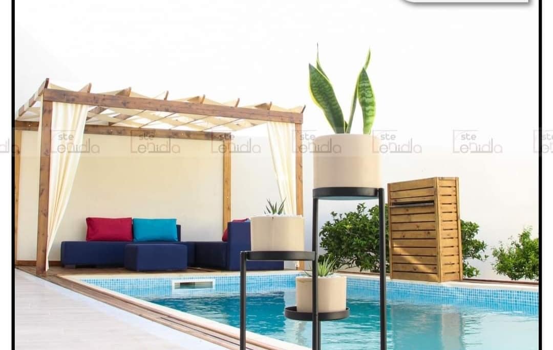A wooden pergola shades a seating area with colorful cushions, positioned next to a swimming pool. Potted plants add greenery, complementing the light blue tiles of the pool. The setting presents a calm atmosphere, ideal for relaxation and enjoyment.