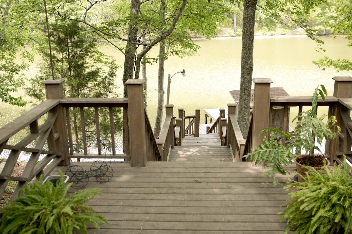 A wooden staircase descends through verdant greenery towards a tranquil lake. The railing is adorned with decorative elements, and ferns are positioned along the steps. Sunlight reflects on the calm water, enhancing the serene outdoor setting.