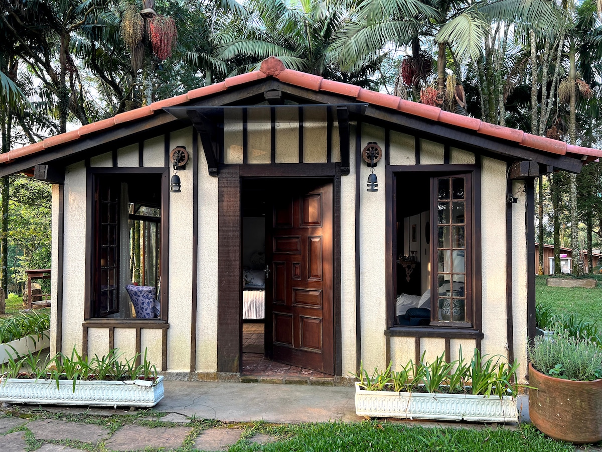 The cozy exterior of a rustic chalet features a warmly lit entrance framed by two windows. Lush greenery and palm trees create a natural backdrop, while the inviting path is lined with planters filled with plants. The distinctive architectural style combines wooden accents with charming details.