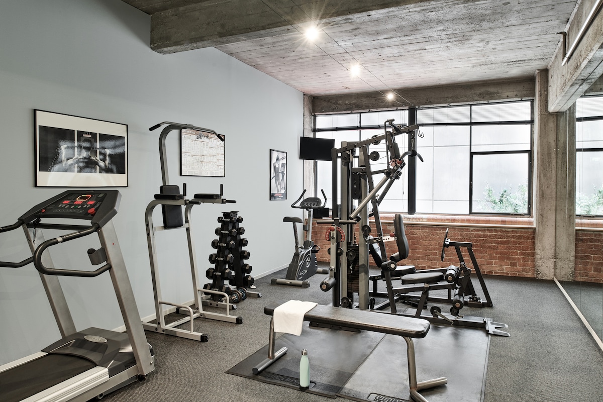 A gym area is equipped with various fitness machines, including a treadmill and weightlifting equipment. A set of dumbbells is arranged on a rack. Large windows allow limited natural light, and concrete walls add an industrial feel to the space.
