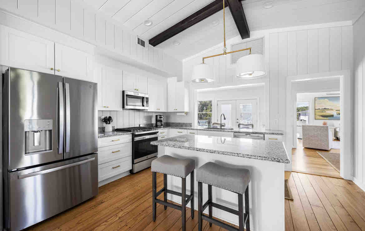 A modern kitchen features crisp white cabinetry and stainless steel appliances, including a large refrigerator and a microwave. A spacious island is complemented by two neutral stools. Natural light fills the area, highlighting the open layout that connects to the adjacent living space.