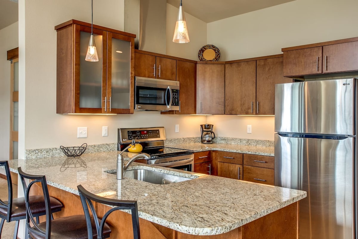 The kitchen showcases warm wood cabinetry and modern stainless-steel appliances. A spacious granite countertop provides seating for two, and well-placed lighting highlights the cooking area. The open layout enhances the functionality of this inviting space.