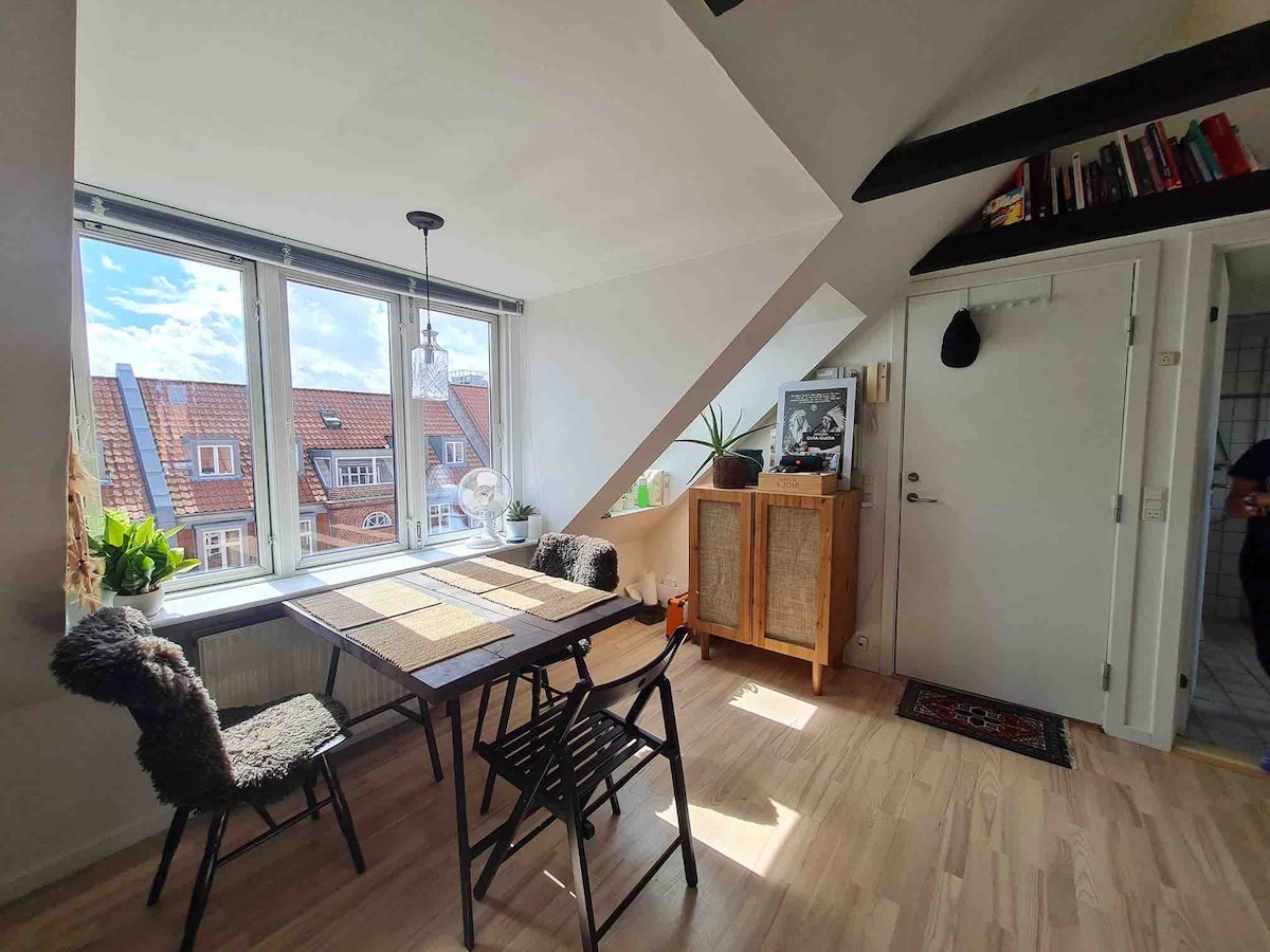 A dining area features a wooden table surrounded by black and gray chairs. A large window allows natural light to illuminate the space, while a small plant sits on the sill. A sideboard provides additional storage, and art decorates the walls.