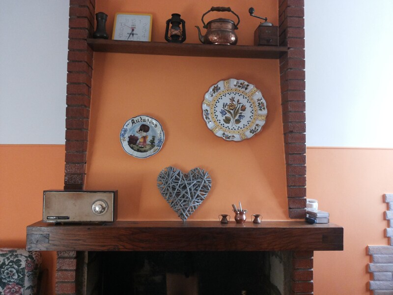 A brick fireplace features a wooden mantel adorned with decorative items, including vintage plates, a heart made of twigs, and a small collection of copper utensils. The wall is painted a warm orange, enhancing the rustic charm of the space.