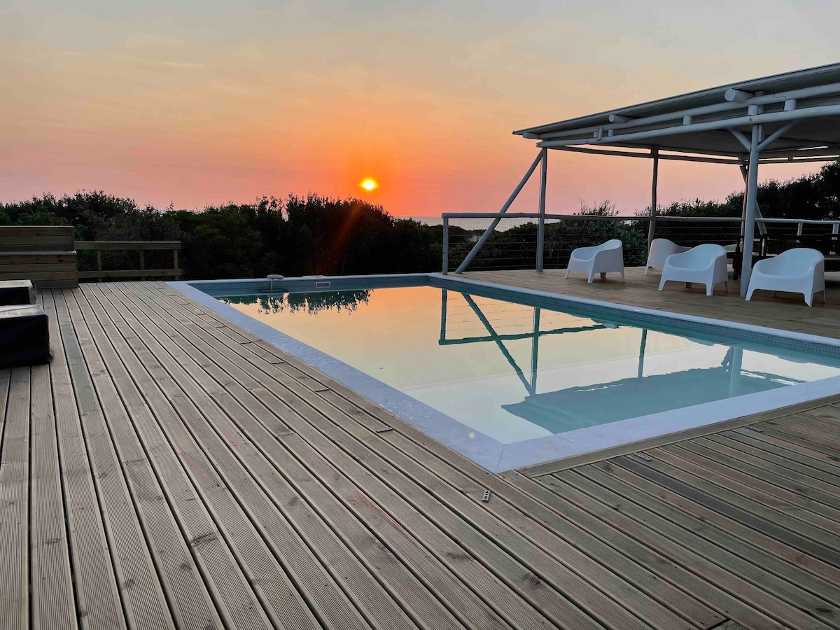 A serene sunset is reflected in a clear private pool, surrounded by a wooden deck. White lounge chairs are positioned near the water, framed by lush greenery in the background. The evening sky transitions from orange to soft purple hues, creating a calm atmosphere.