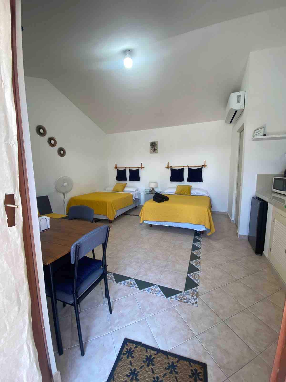 The room features two spacious beds covered in yellow bedspreads. A small dining table with chairs is positioned nearby, alongside a fan. A microwave is situated in the kitchenette area. Natural light fills the space through a window, enhancing the simple decor.