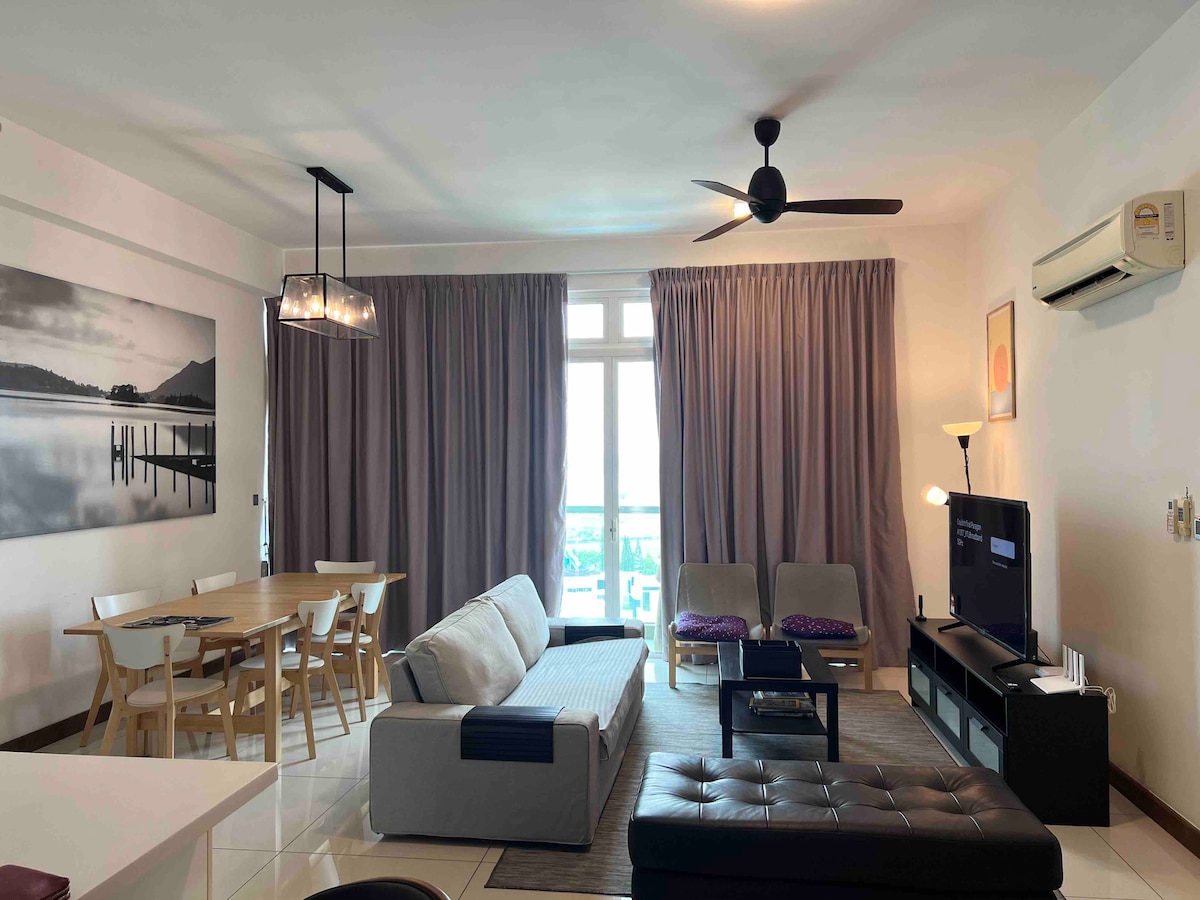 A spacious living area features large windows with natural light and soft drapes, providing a view of the surroundings. A comfortable sectional sofa is positioned in front of a TV, accompanied by two armchairs. A wooden dining table with chairs rounds out the cozy atmosphere.