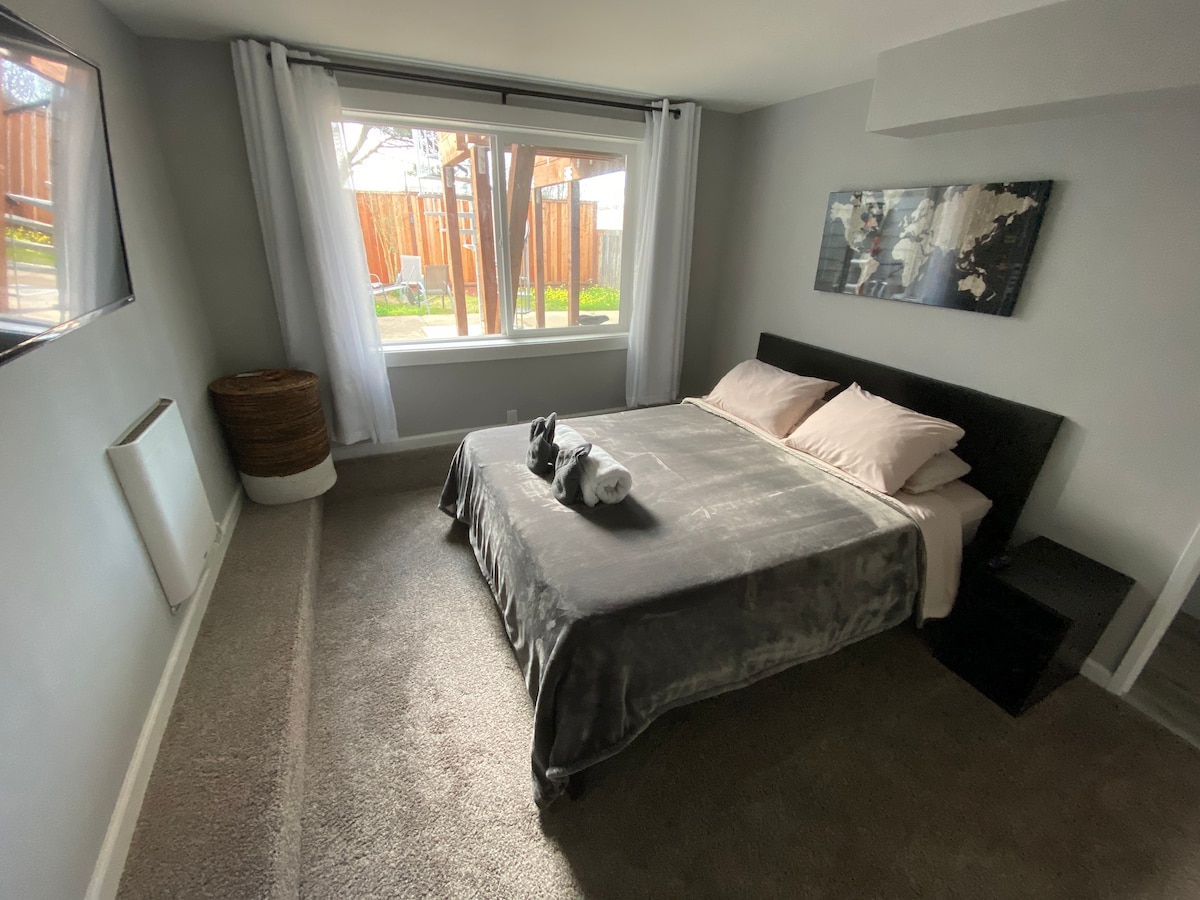 The bedroom features a spacious bed dressed in soft bedding, complemented by a bedside table. A large window allows natural light to flood the space, revealing a view of a backyard garden. A flat-screen TV is mounted on the wall opposite the bed.