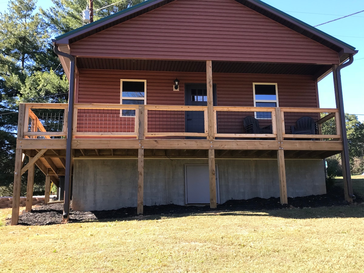 The exterior of the house features a sturdy wooden deck with seating options, set against a backdrop of a well-maintained lawn. The warm-colored wooden siding gives the home a welcoming appearance, complemented by nearby trees and a clear sky.
