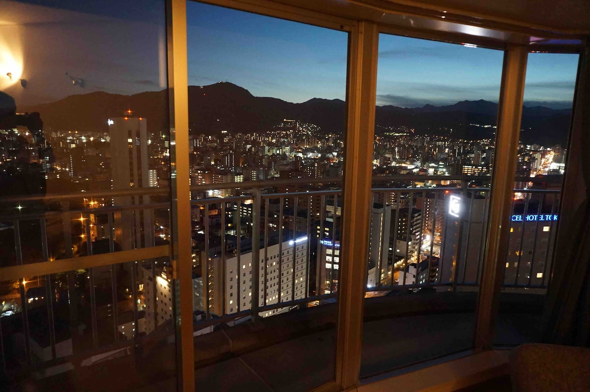 A panoramic view of Sapporo is presented through large glass windows, showcasing the city lights against a dusky sky. Mountains are faintly visible in the background, adding to the serene atmosphere of the scene.