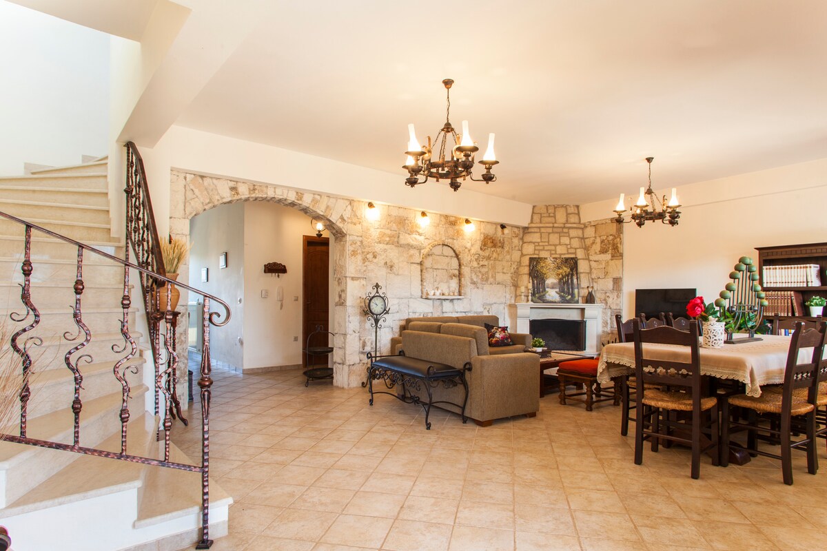 An open-plan living and dining area features traditional stone walls and elegant chandeliers. A comfortable sofa and dining table are positioned to create inviting spaces for relaxation and meals. A staircase is visible in the background, leading to the upper floor.