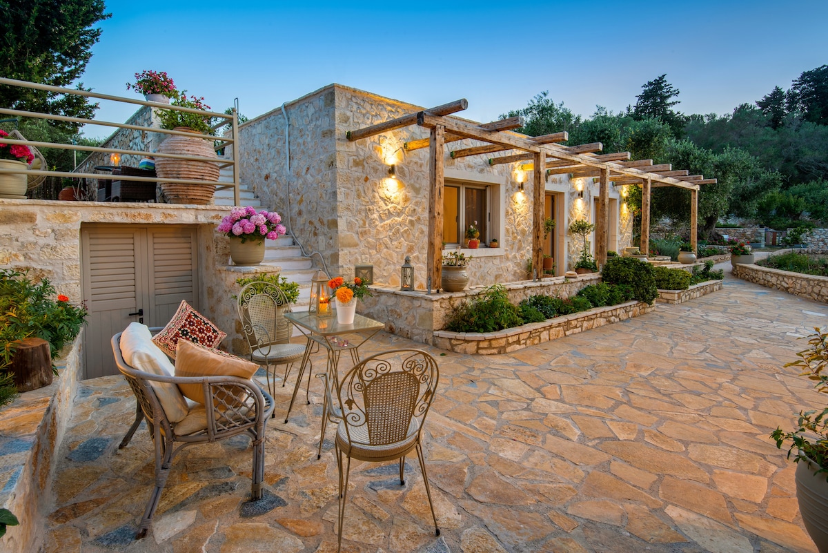 A charming stone patio is lined with potted flowers and features a small seating area. A table and chairs are positioned at the entrance, surrounded by lush greenery and a peaceful atmosphere. The exterior of the house is well-lit, highlighting its rustic design.