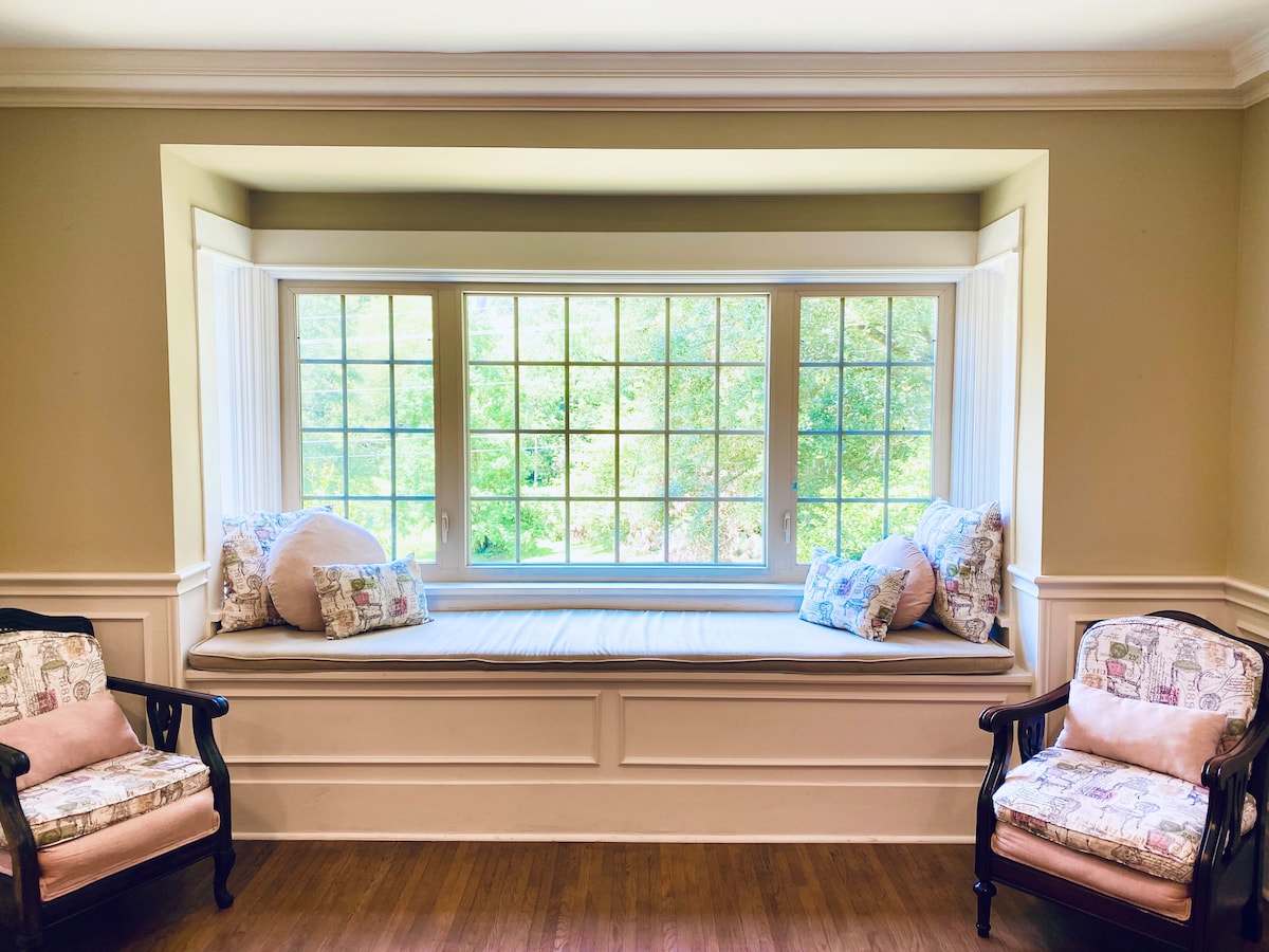 A cozy window seat is framed by large windows, inviting natural light to fill the space. Soft cushions are arranged on the bench, complemented by two matching chairs positioned nearby. The view reveals a lush garden, enhancing the tranquil atmosphere.