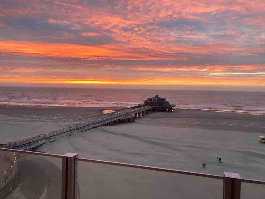 A vibrant sunset is displayed over the ocean, with hues of orange and pink reflecting on the water. A wooden pier extends into the sea, while small figures are seen walking along the beach. The view is framed by a glass railing.