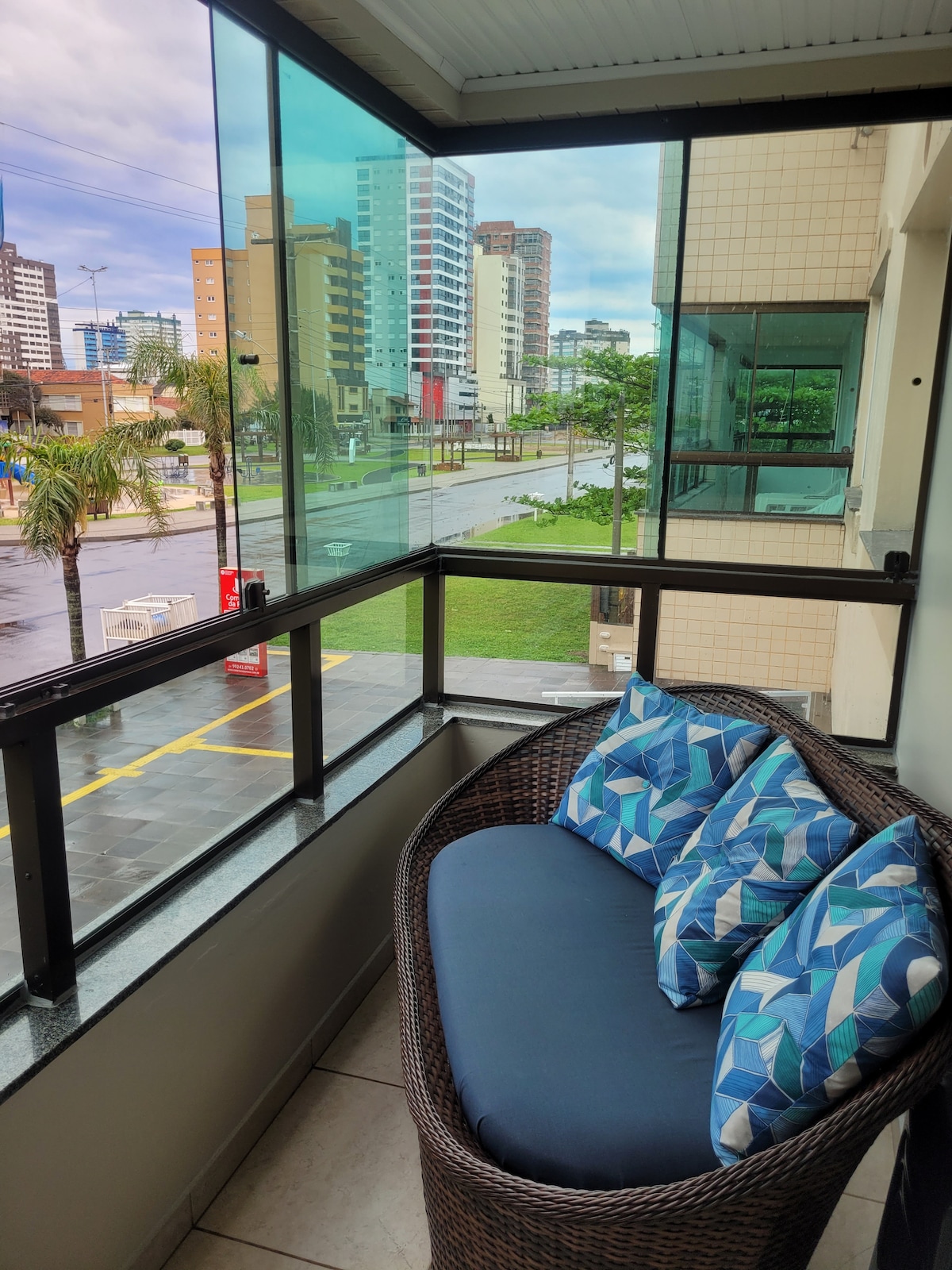 A balcony area features a comfortable wicker sofa adorned with patterned cushions. Large glass panels provide a view of the surrounding cityscape, with buildings and greenery visible in the distance. The space offers a quiet retreat with ample natural light.