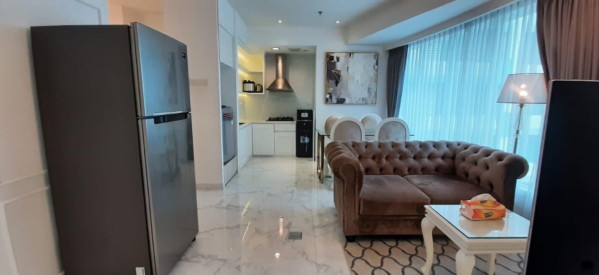 A modern living area features a comfortable brown chesterfield sofa facing a glass coffee table. An open kitchen showcases sleek white cabinetry and high-end appliances. Natural light pours in through large floor-to-ceiling windows, illuminating the polished marble floor.