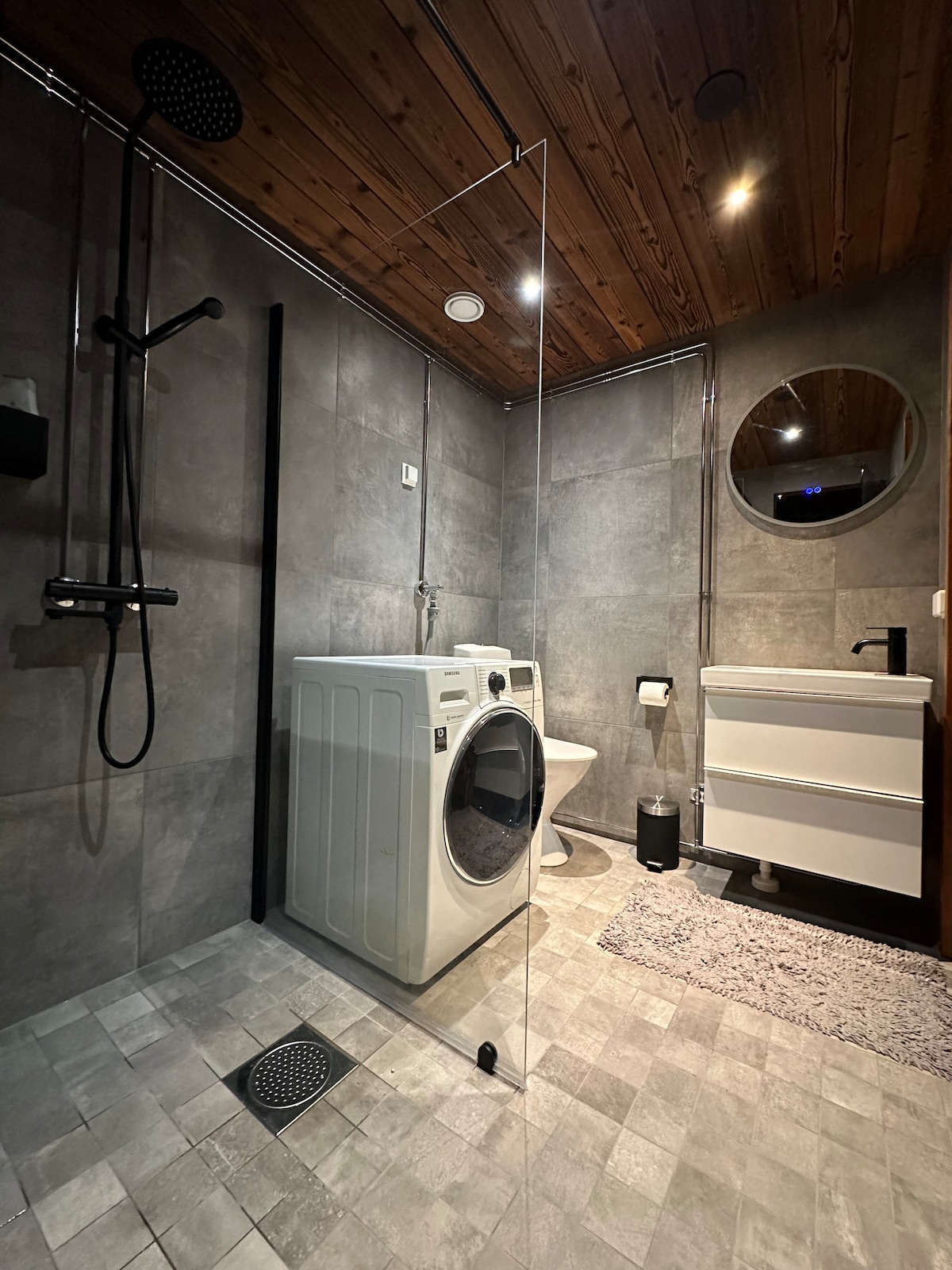 The modern bathroom features a glass-enclosed shower with a rainfall showerhead, a sleek vanitory with a round mirror, and a washing machine. Neutral-toned tiles cover the floor, while warm wooden paneling accents the ceiling, enhancing the contemporary design.