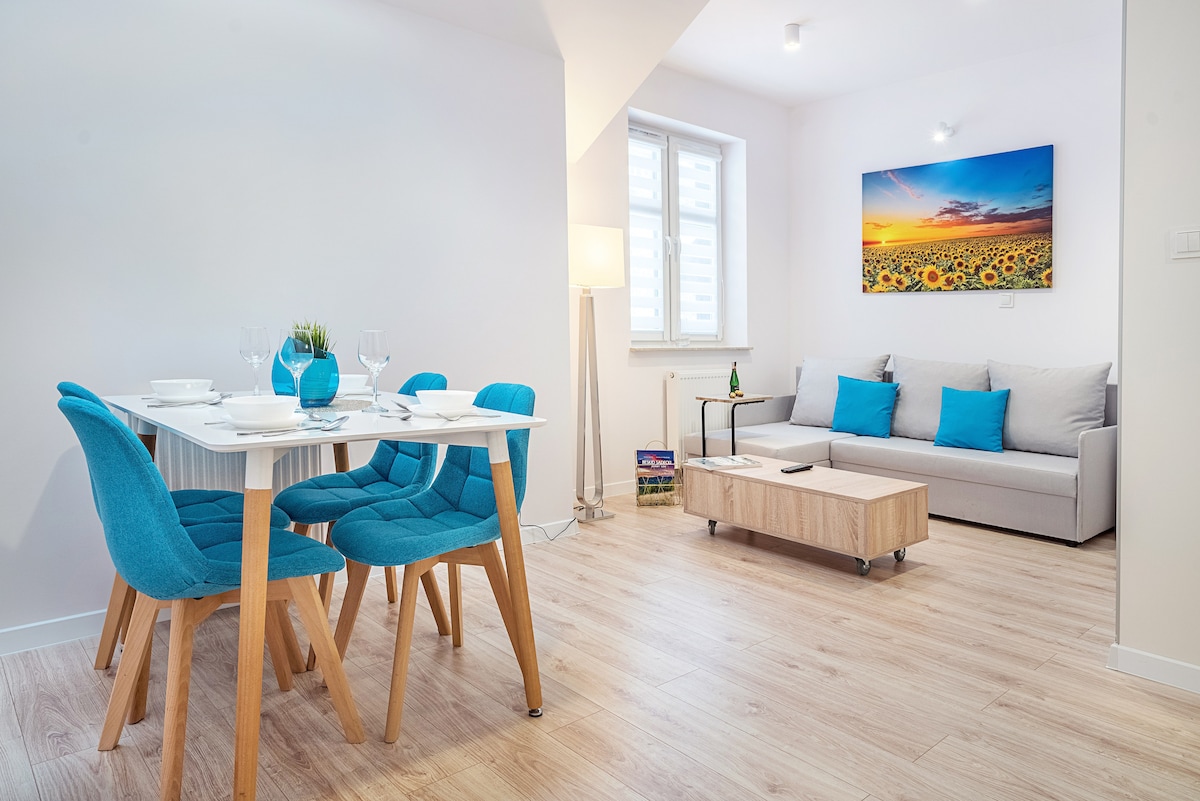 A modern dining area features a white table surrounded by blue chairs. Adjacent, a light gray sofa sits across from a coffee table. Sunflower artwork adorns the wall, and natural light is present through a window, enhancing the spacious, airy feel of the room.