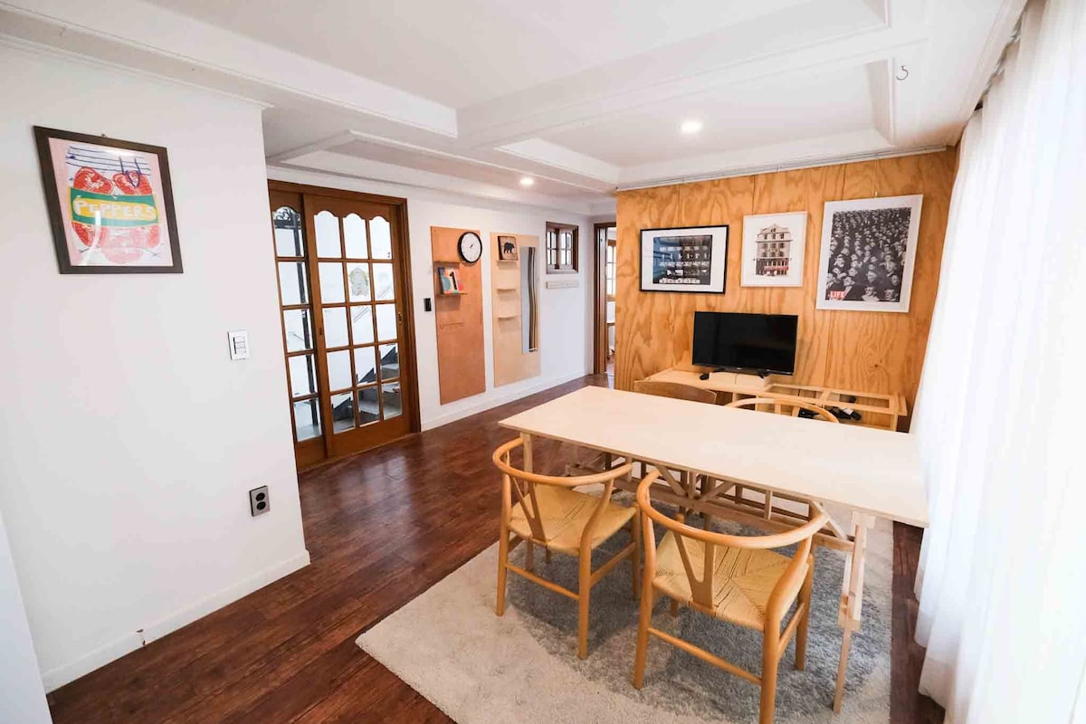 A bright living area features a wooden table surrounded by four elegant chairs on a soft rug. A mounted television is positioned against a warm wooden wall, while light streams in through large windows covered by sheer curtains. Decorative art pieces add character to the space.