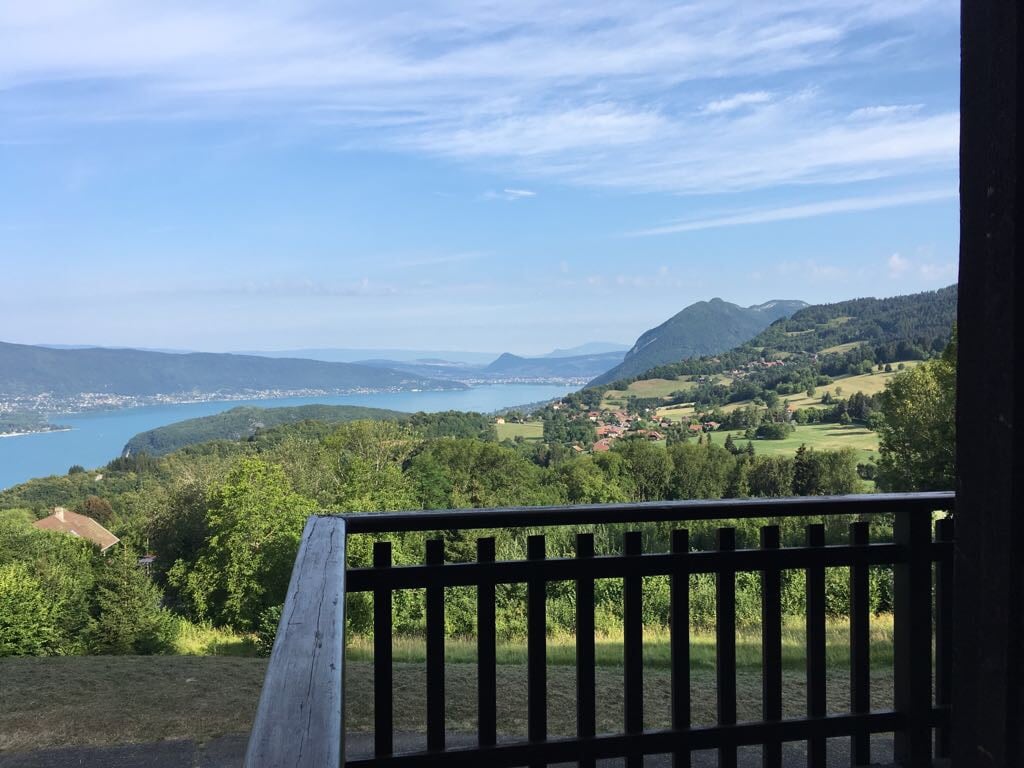 A scenic view of Lake Annecy is displayed from a balcony, featuring lush green hills and distant mountains. The expansive body of water reflects the sky, creating a serene landscape. A wooden railing frames the view, providing a sense of openness and connection to the outdoor environment.