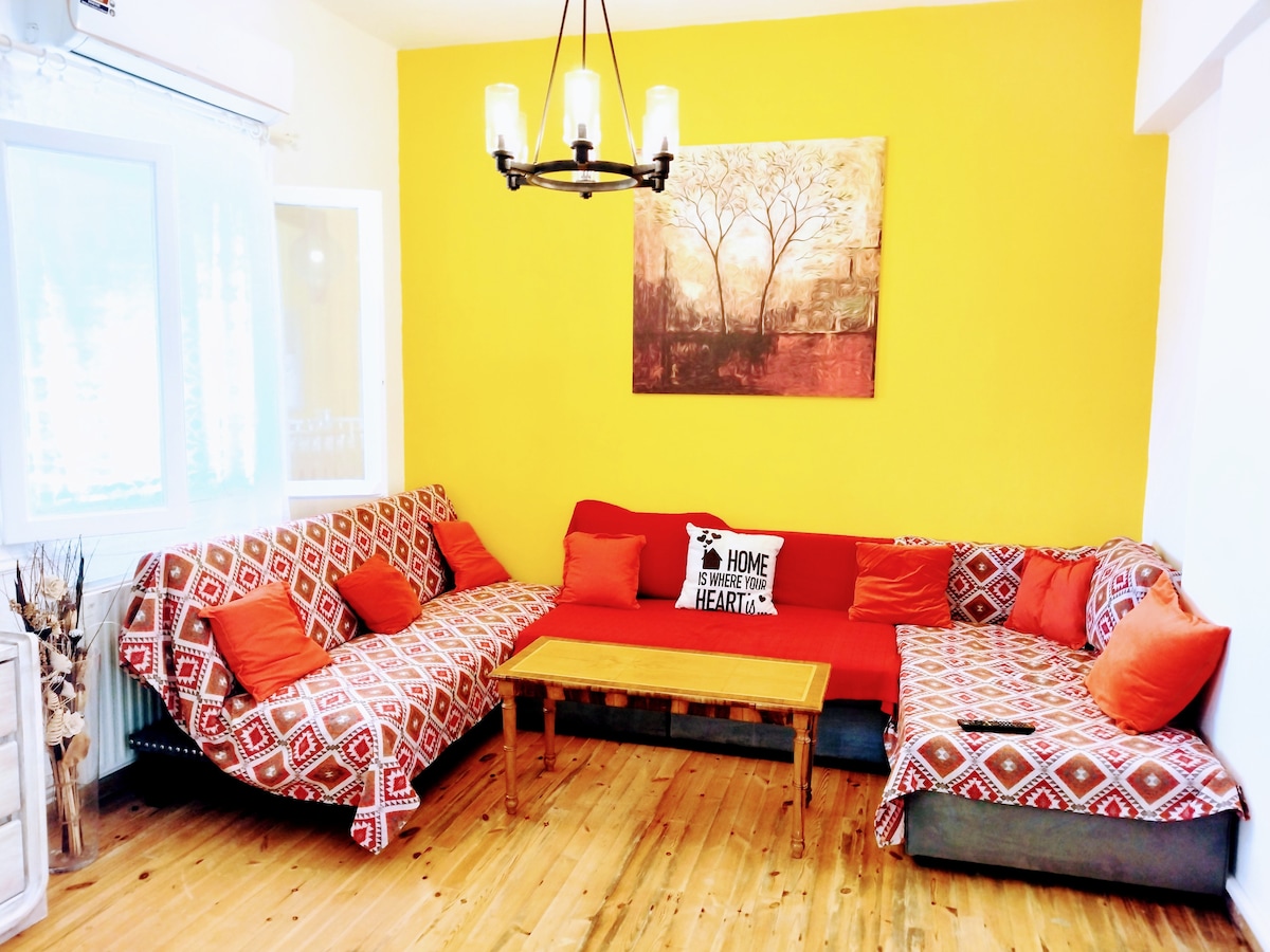 A cozy living area features a large corner sofa adorned with patterned throws and vibrant orange cushions. A wooden coffee table occupies the center, and a painting hangs on the bright yellow wall, creating a welcoming space. Natural light filters in through the nearby window.