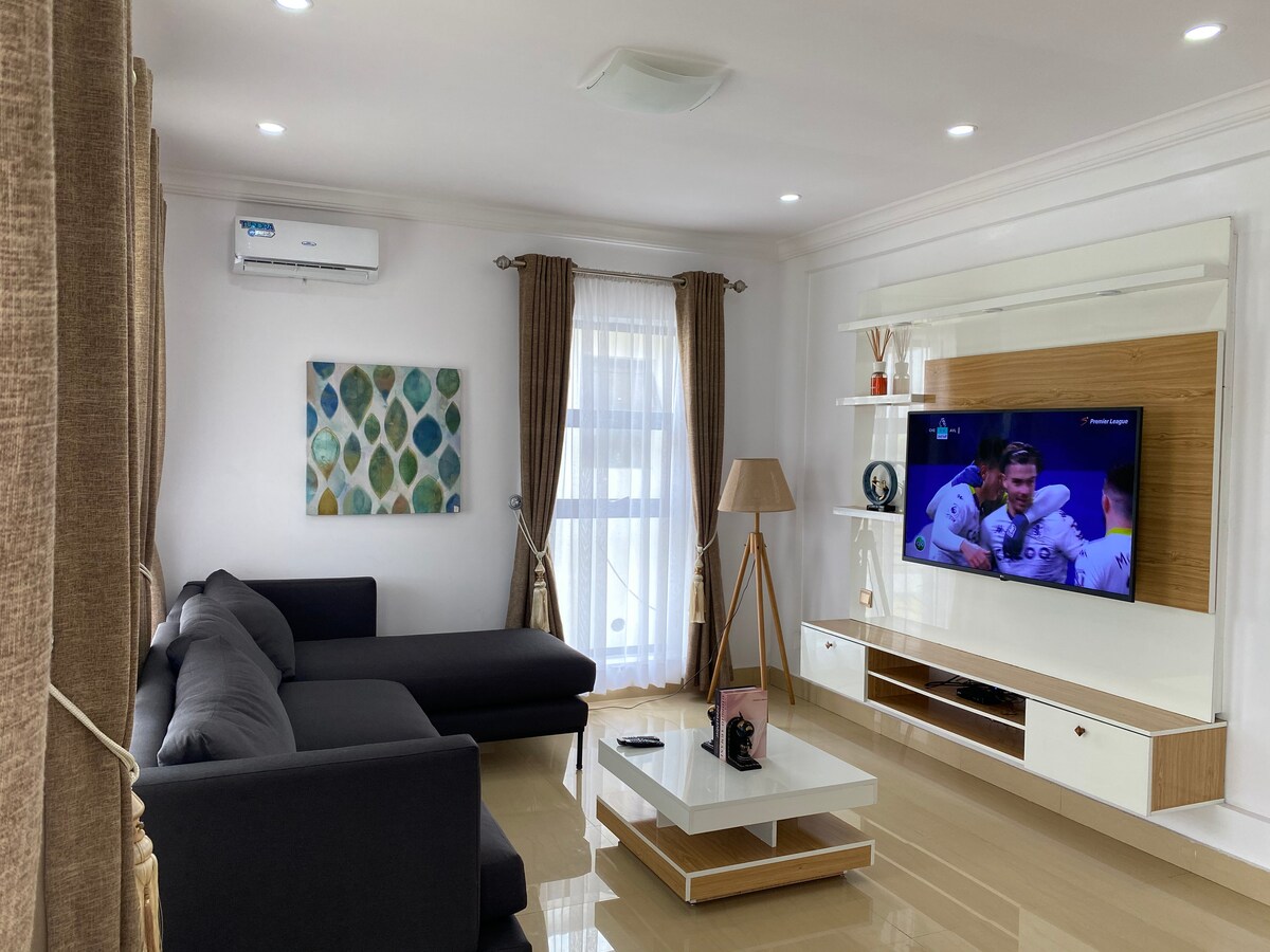 A modern living area features a dark gray sectional sofa positioned beside a sleek white entertainment unit. A flat-screen television is mounted on the wall, while natural light enters through large windows covered by light curtains. A decorative lamp and artwork add subtle detail to the space.