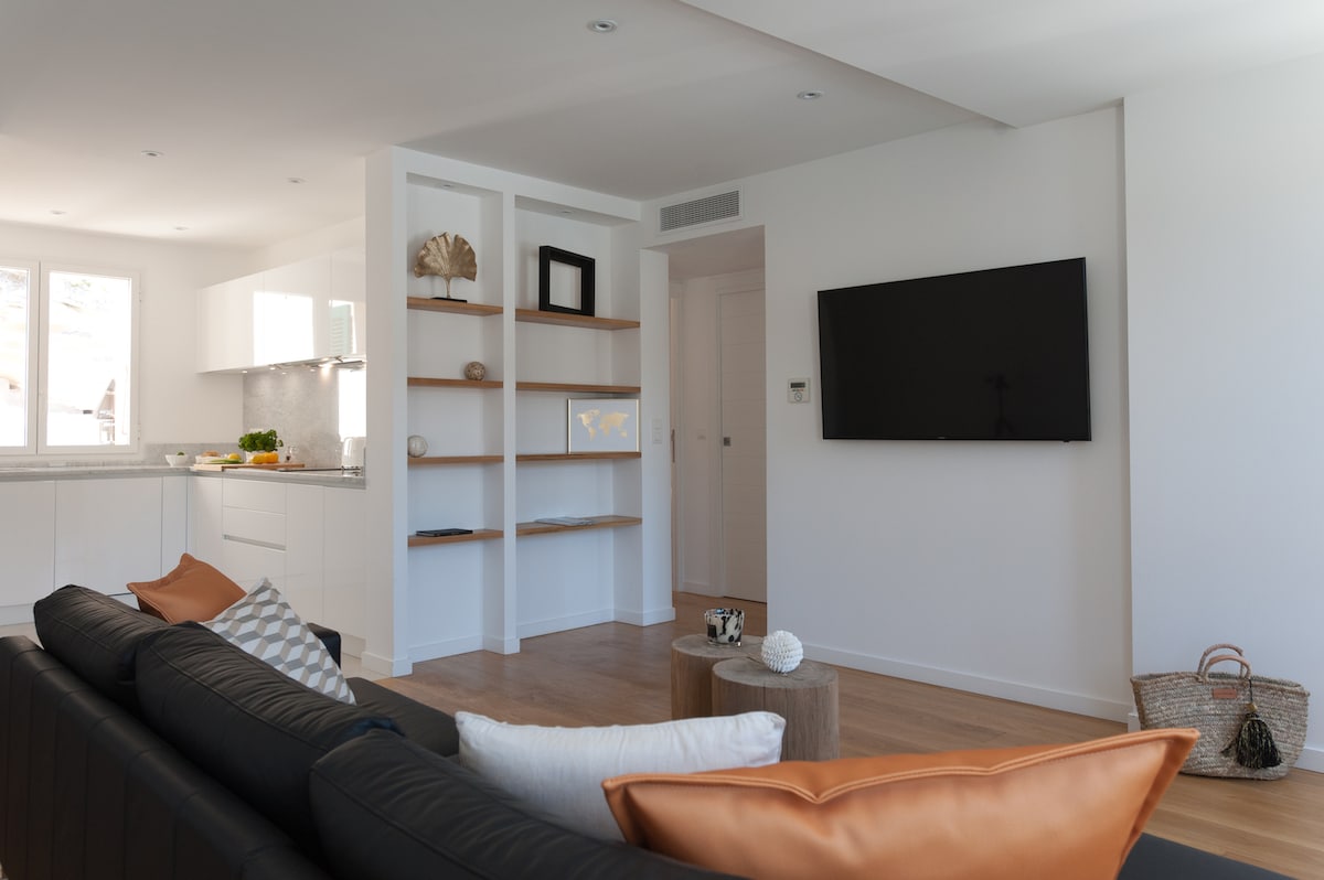 A contemporary living area features a sleek black sofa adorned with decorative cushions. A minimalist wooden coffee table is positioned in front of the sofa, while a large flat-screen TV is mounted on the wall. Light streams in from the nearby windows, illuminating the space.