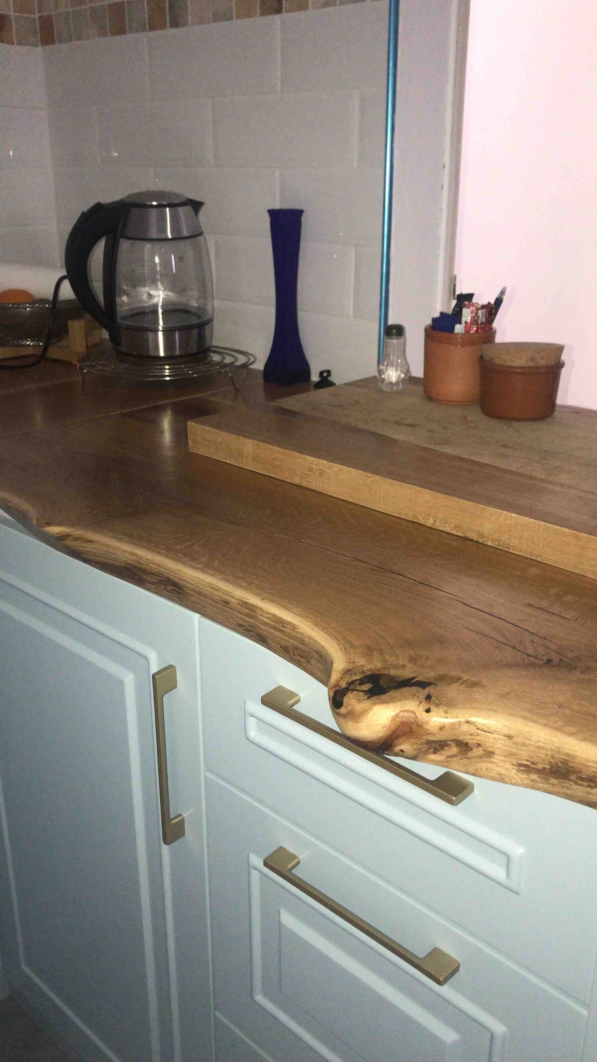 A kitchen counter features a smooth, natural wood surface with unique grain patterns, complemented by light blue cabinetry. A kettle and other kitchen items are arranged on the counter, which also includes a wooden cutting board and various small containers for utensils.