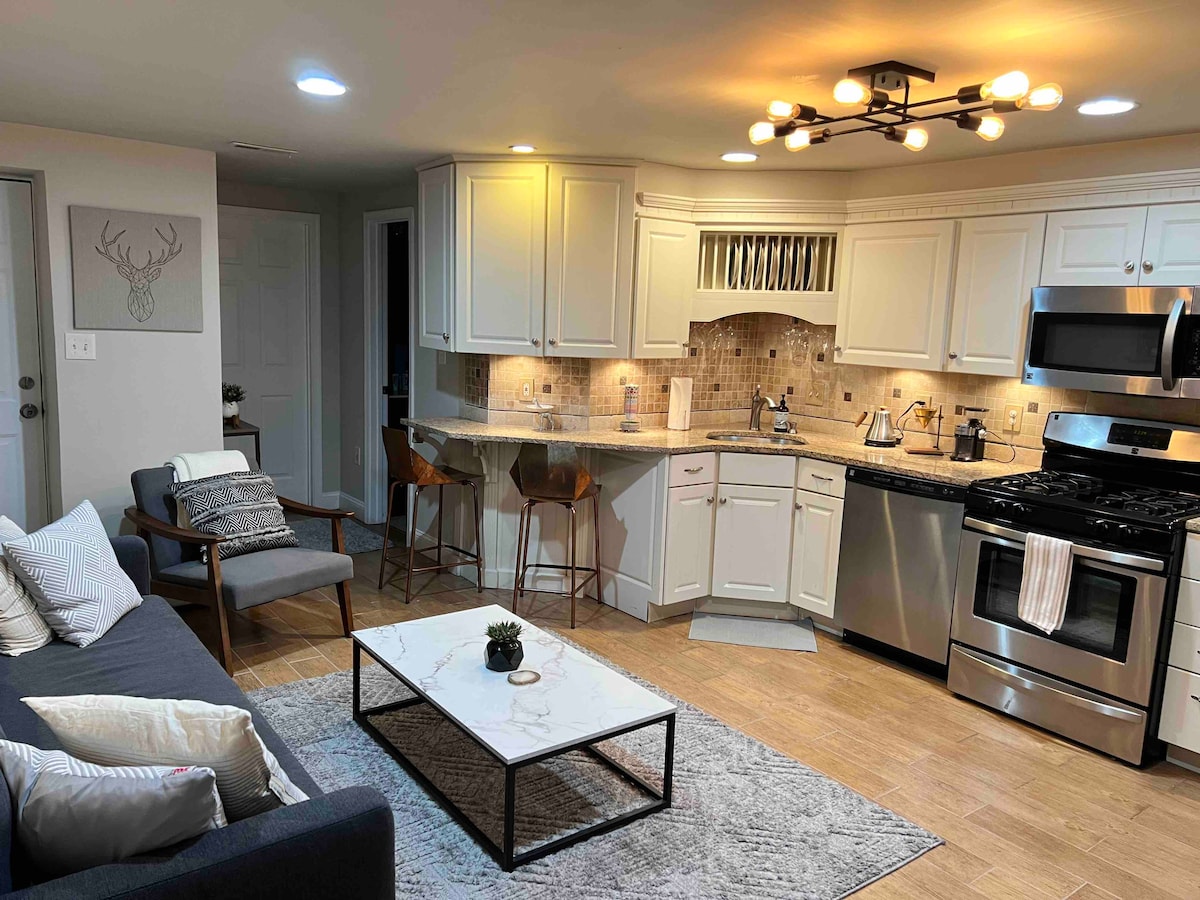 The kitchen and living area are shown in an open layout, featuring modern appliances and a spacious countertop. Two bar stools provide seating at the counter. A comfortable sofa is positioned near a coffee table on a light area rug, illuminated by warm overhead lighting.