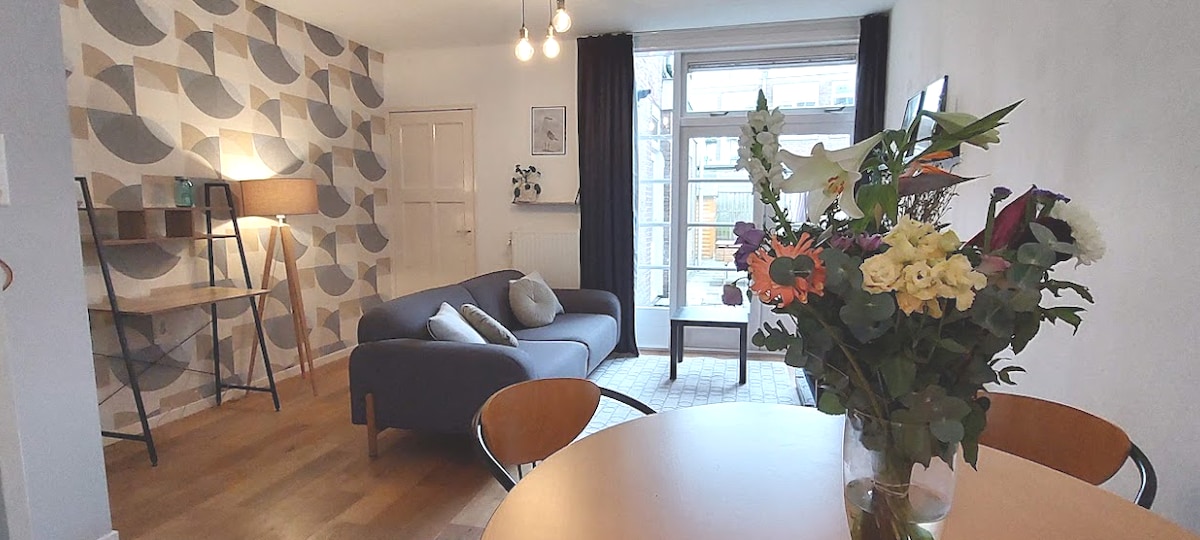 A cozy living space features a soft gray sofa, complemented by a small side table and a warm floor lamp. A dining table with two chairs is positioned nearby. Large windows allow natural light to illuminate the room, with decorative wall patterns adding visual interest.