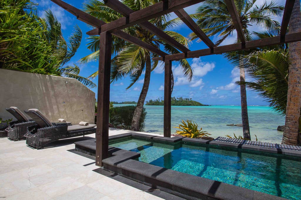 A private outdoor area features a refreshing plunge pool surrounded by tropical palm trees. Chaise lounges are positioned nearby for relaxation, with views of the azure lagoon visible in the background under a clear blue sky.